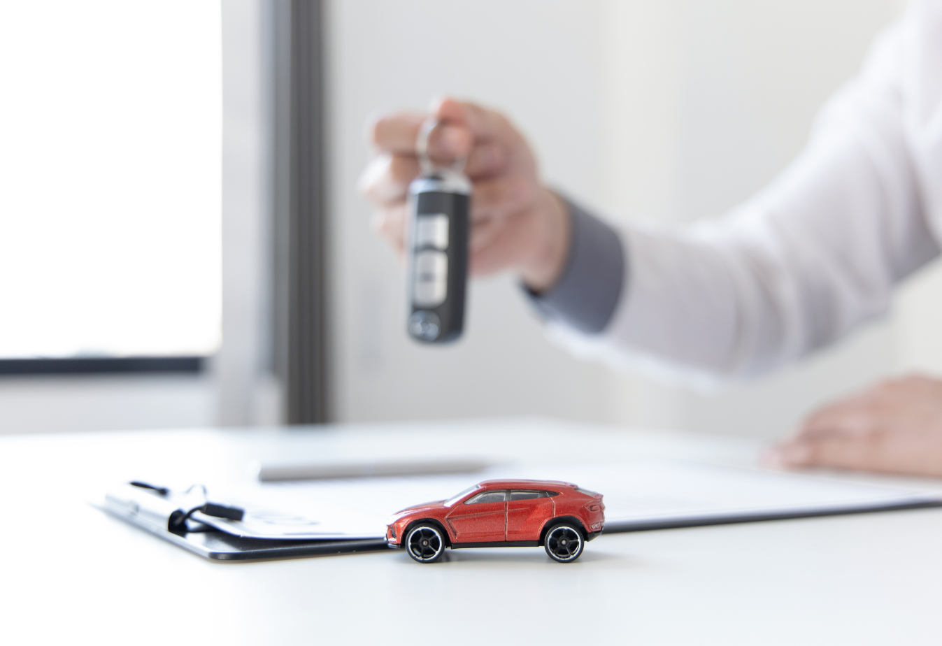 a person holding car keys over a clipboard with a small red toy car in the foreground